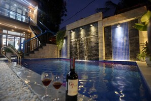 A swimming pool with a waterfall feature, a bottle of wine, and two glasses on the poolside.