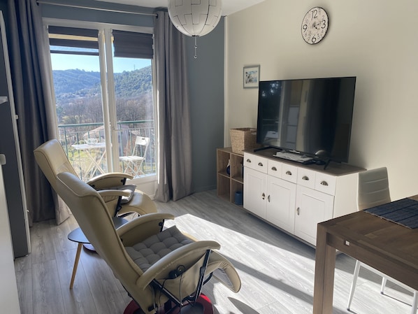 A modern living room with a large TV, a comfortable reclining chair, a dining table, and a view of the outdoors through a large window.