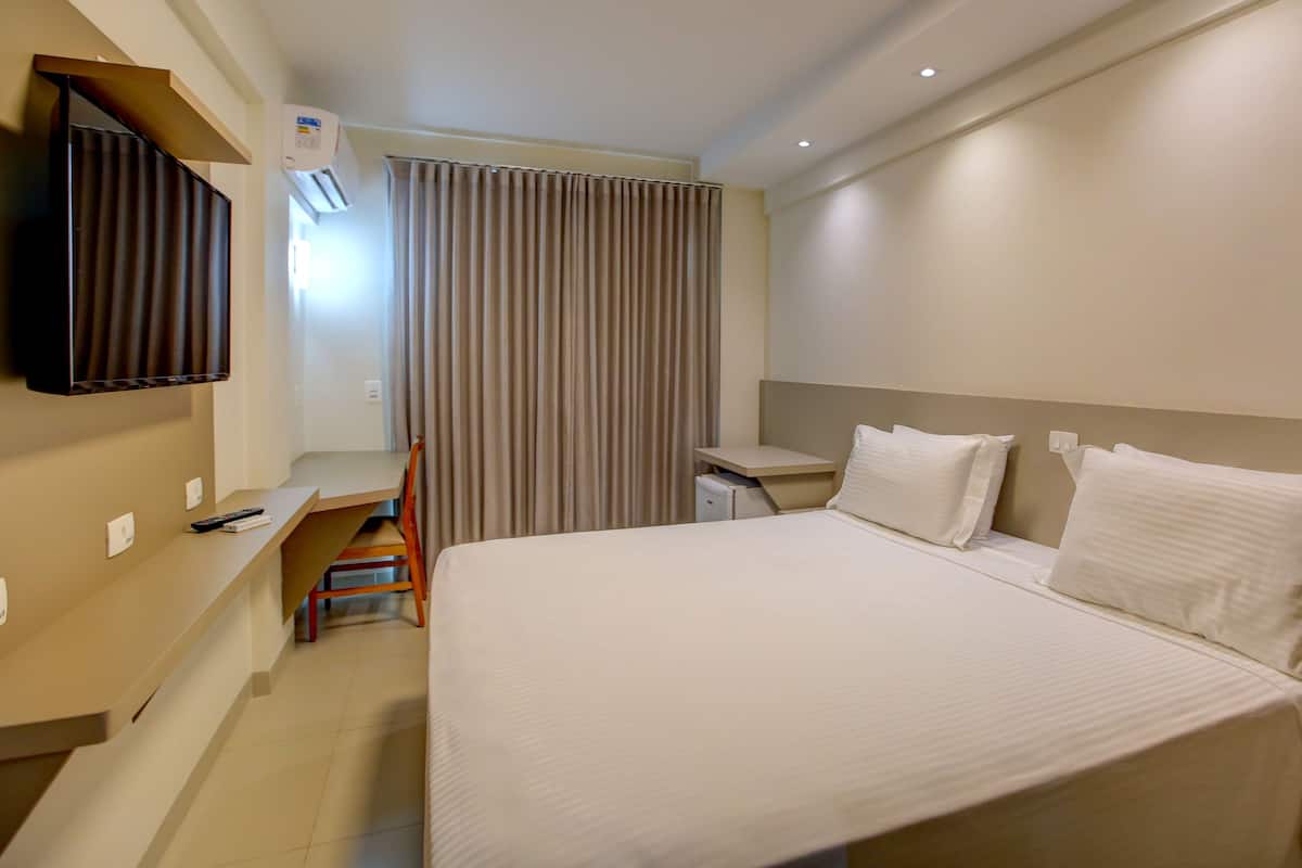 A hotel room with a bed, a desk, a chair, a TV, and an air conditioning unit.