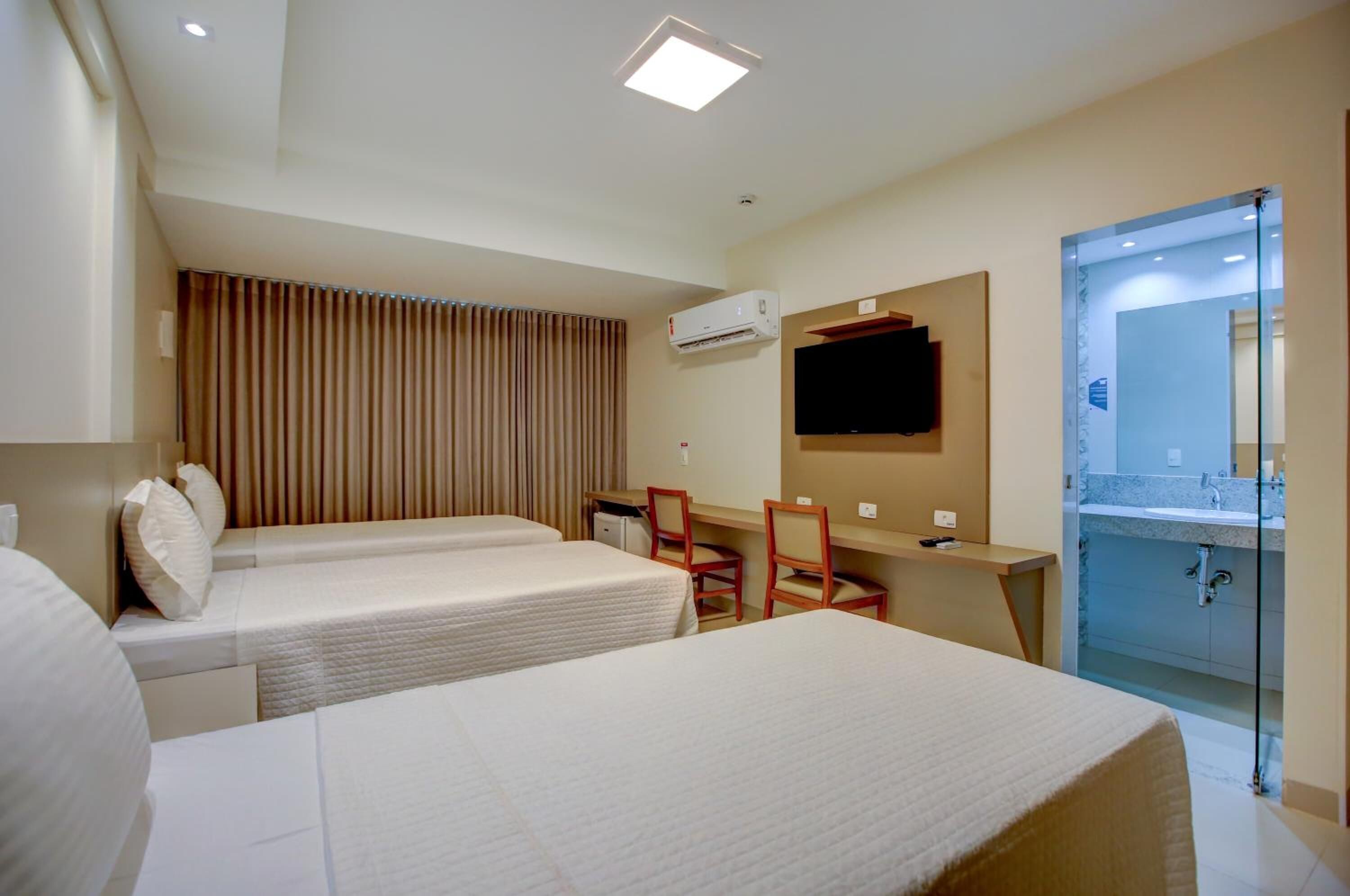 Habitación de hotel con dos camas, un escritorio, una silla, un televisor, un equipo de aire acondicionado y un baño con separador de vidrio.