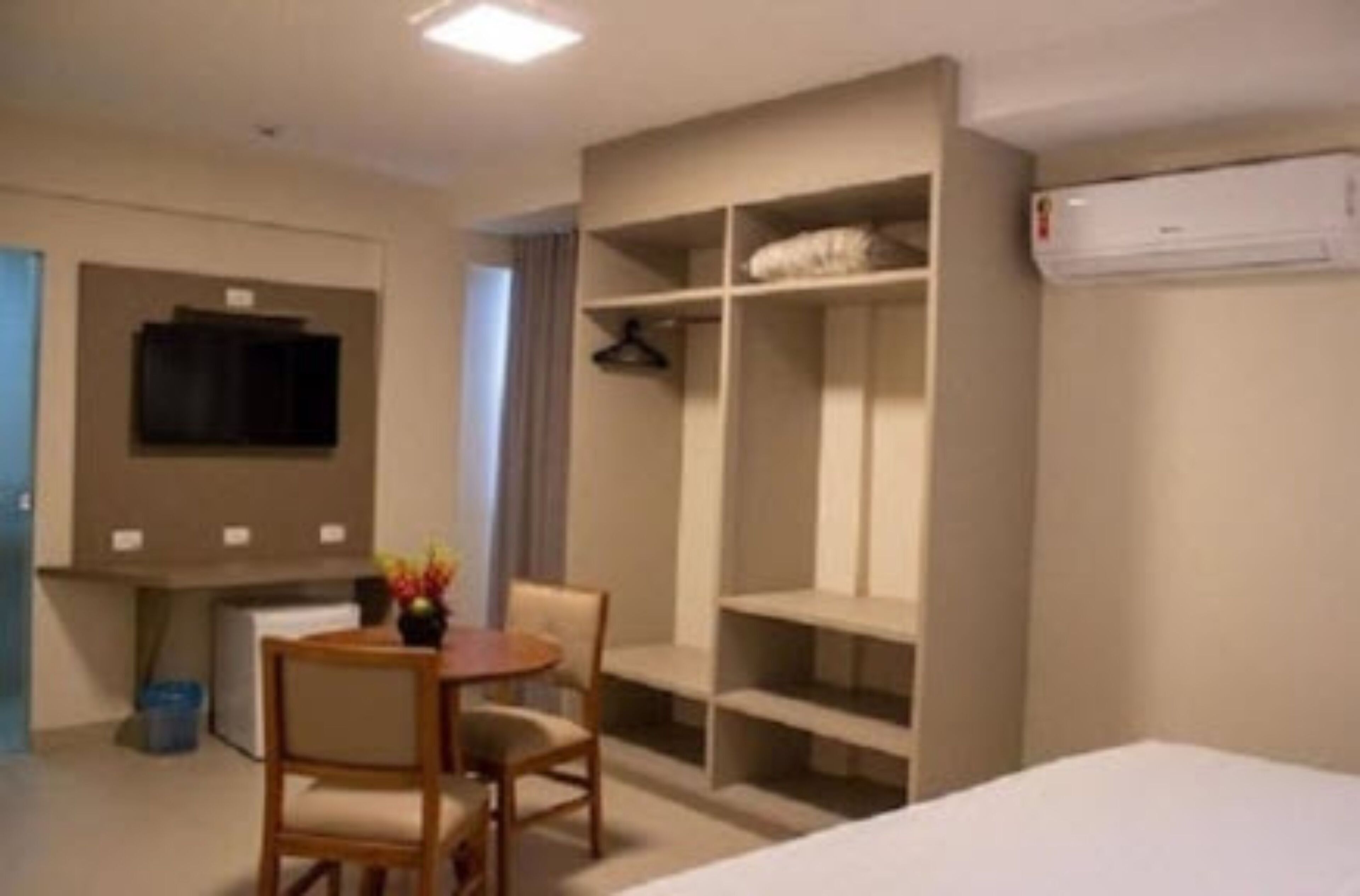 A compact hotel room with a bed, a small table with chairs, a wardrobe, a TV, and an air conditioning unit.