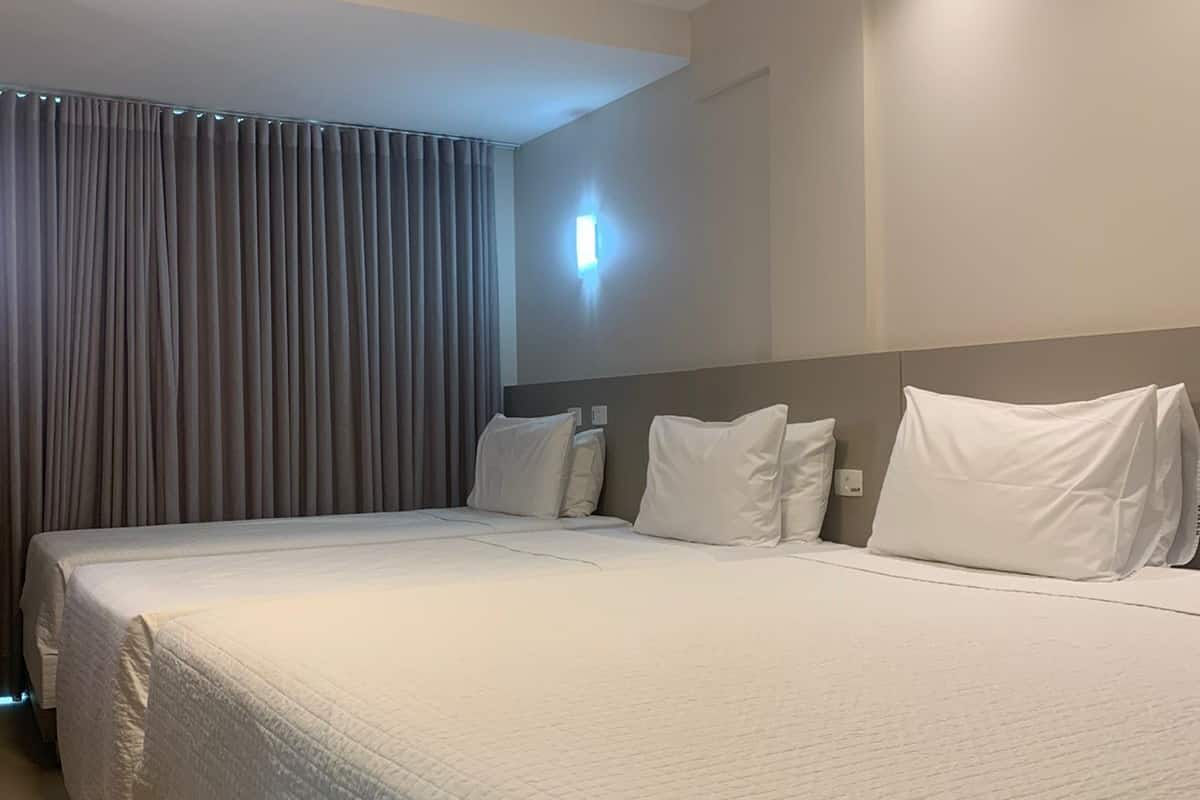 A hotel room with two beds, a large window with curtains, a wall-mounted light, and a ceiling with recessed lighting.