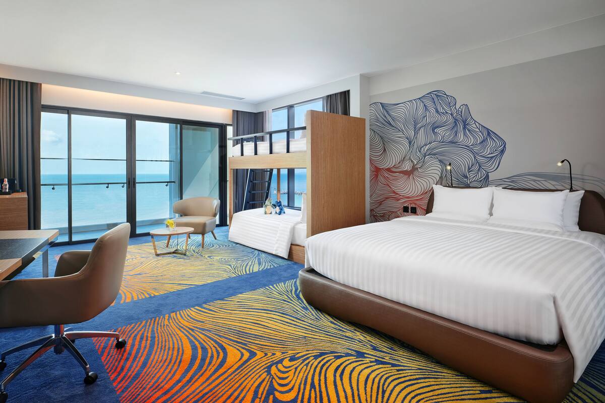 Ein Hotelzimmer mit einem großen Bett, einem Schreibtisch, einem Stuhl und Meerblick durch die Fenster.