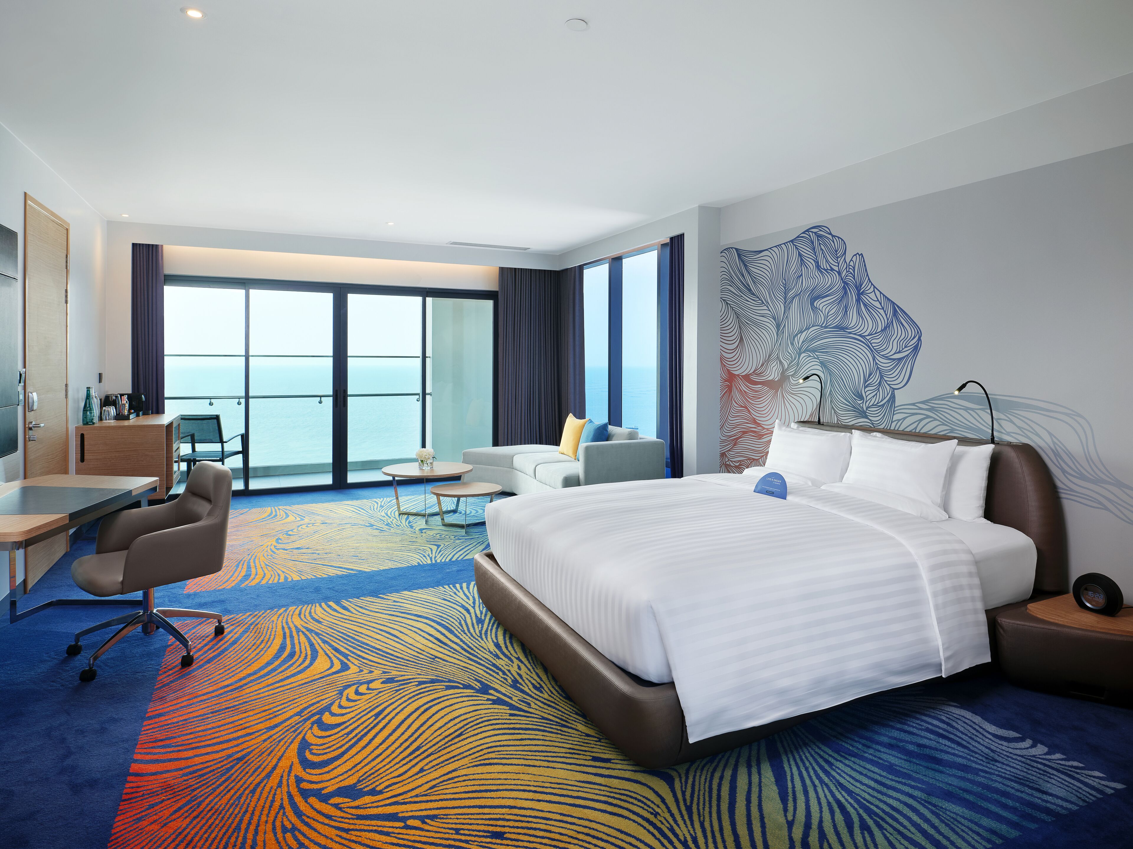 A hotel room with a large bed, a desk, a chair, a small table, and a view of the ocean through large windows.