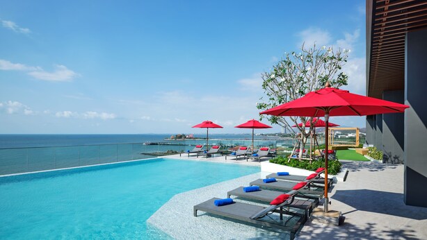 Ein Poolbereich mit Liegestühlen und roten Sonnenschirmen mit Blick auf das klare, blaue Meer.
