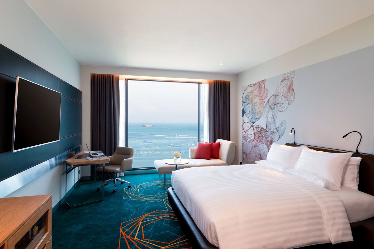 Ein Hotelzimmer mit einem großen Bett, einem Schreibtisch mit einem Laptop, einem Stuhl, einem kleinen Tischchen mit einer Vase und Meerblick durch ein großes Fenster.
