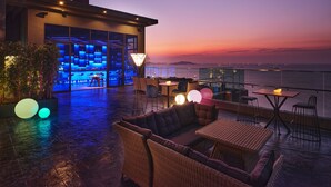 Eine Dachterrasse mit Blick auf das Meer bei Sonnenuntergang, ausgestattet mit Korbmöbeln und stimmungsvoller Beleuchtung.