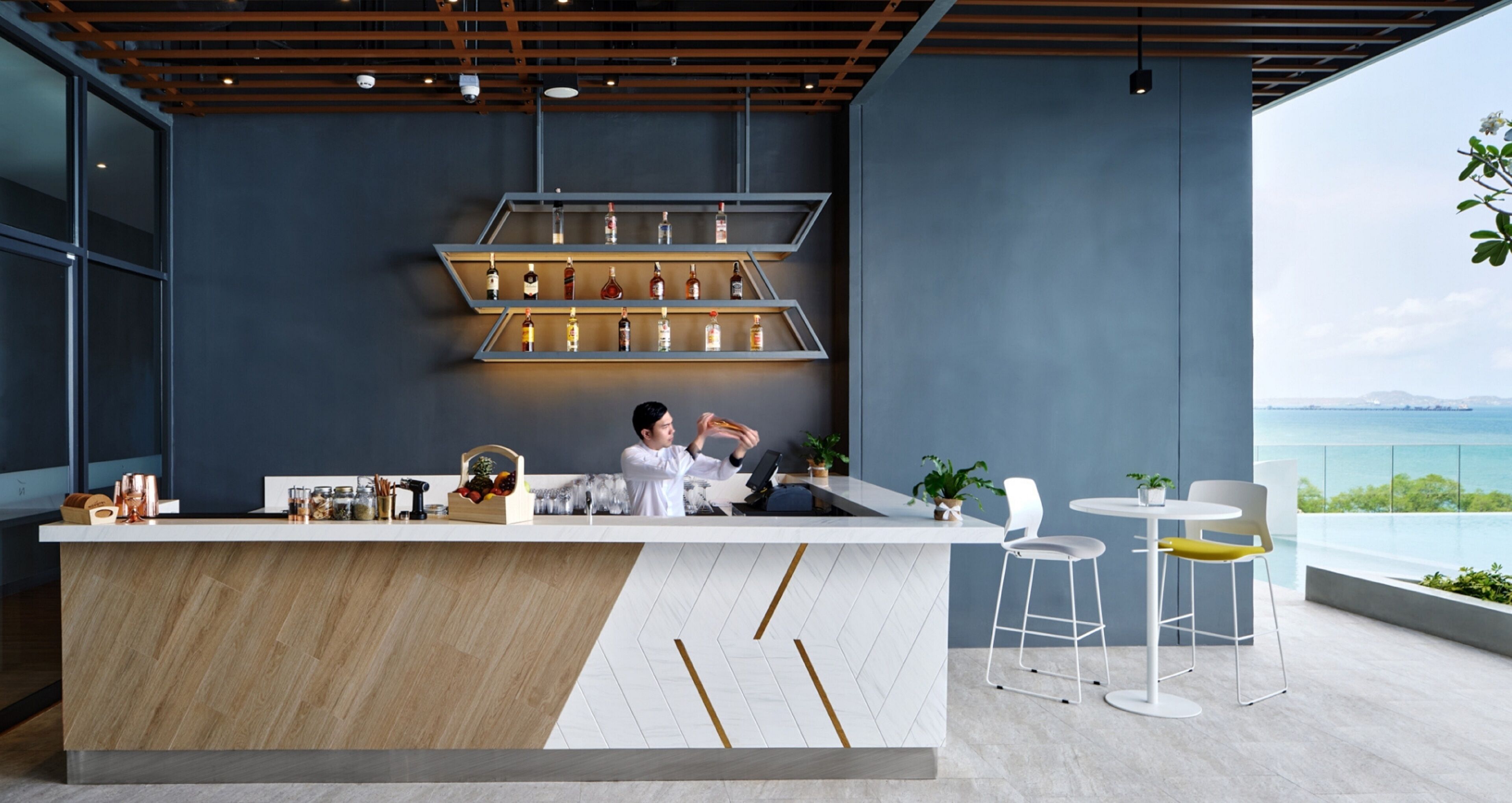 Un espace bar moderne avec un comptoir en bois, des étagères remplies de bouteilles et une vue sur la mer.