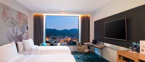 Habitación de hotel con dos camas, un escritorio, una silla, un televisor y vista a la ciudad al anochecer.