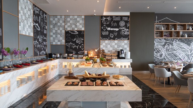 A modern restaurant with a marble bar, various food stations, and a seating area with chairs and tables. - Novotel Marina Sriracha (Si Racha)