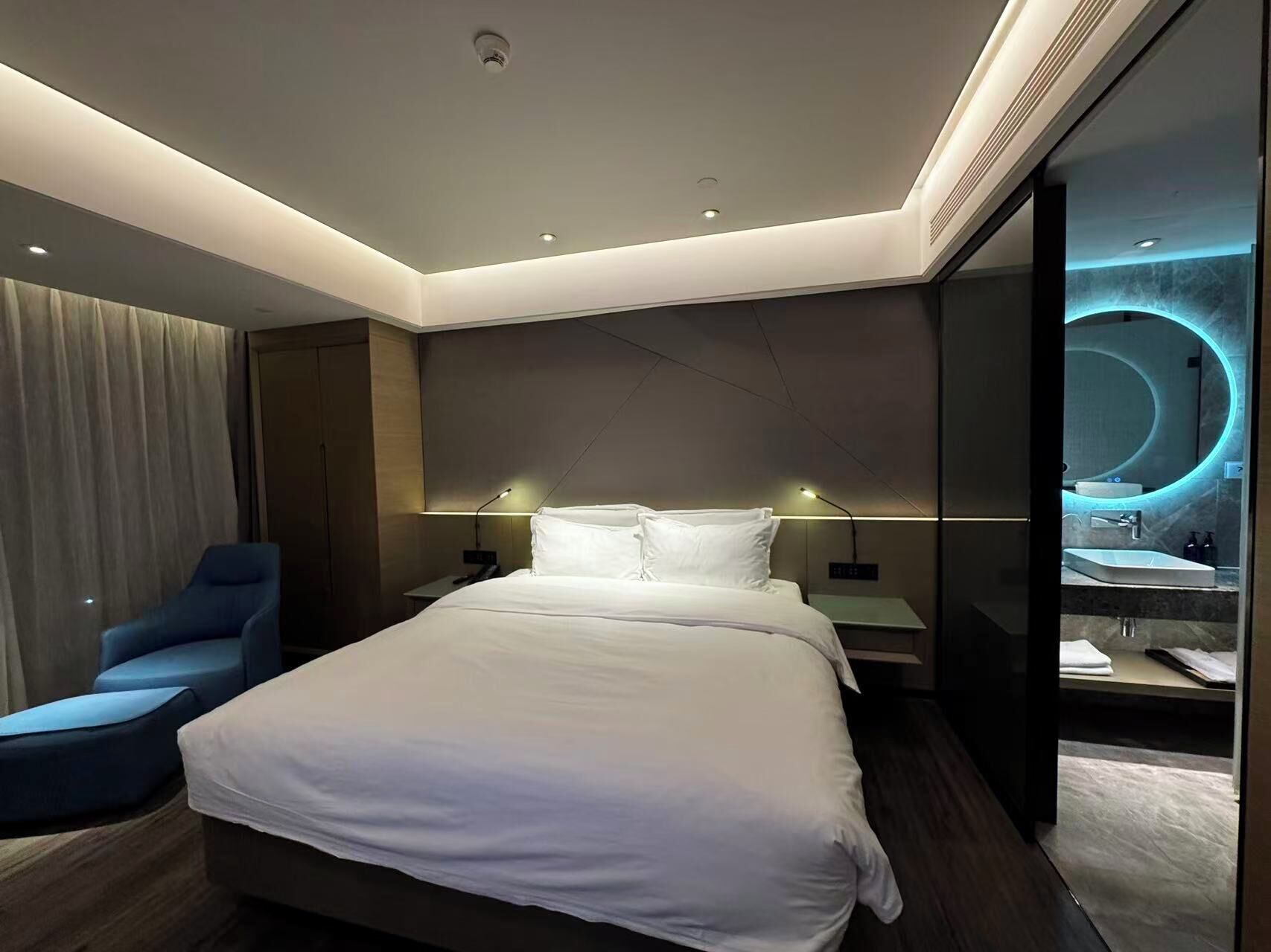 A modern hotel room with a large bed, a desk, a chair, and a bathroom with a circular mirror.