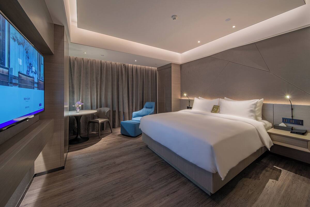 A modern hotel room with a large bed, a desk, a blue chair, and a television on the wall.