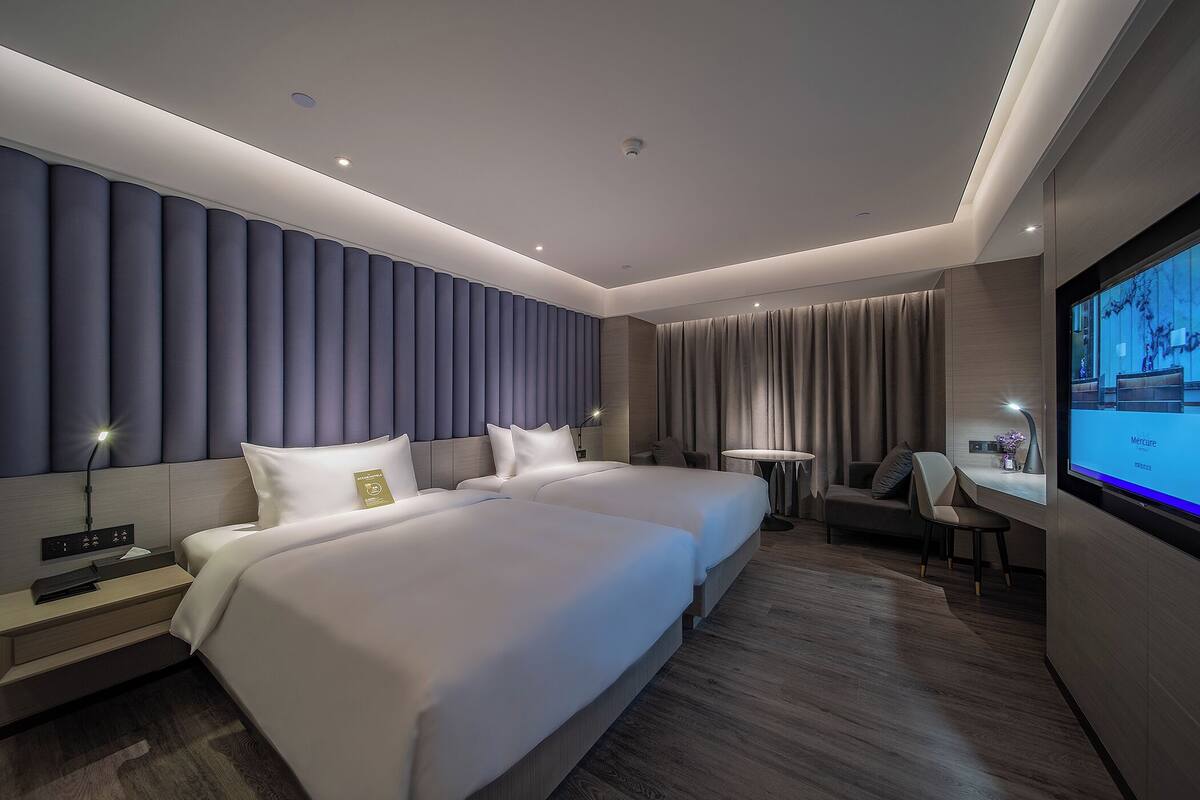 A modern hotel room with a large bed, bedside tables, a desk, a chair, a TV, and a view of the city.