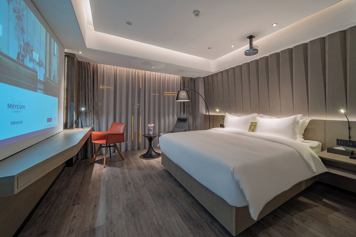 A modern hotel room with a large bed, a desk, a chair, and a TV screen displaying hotel information.