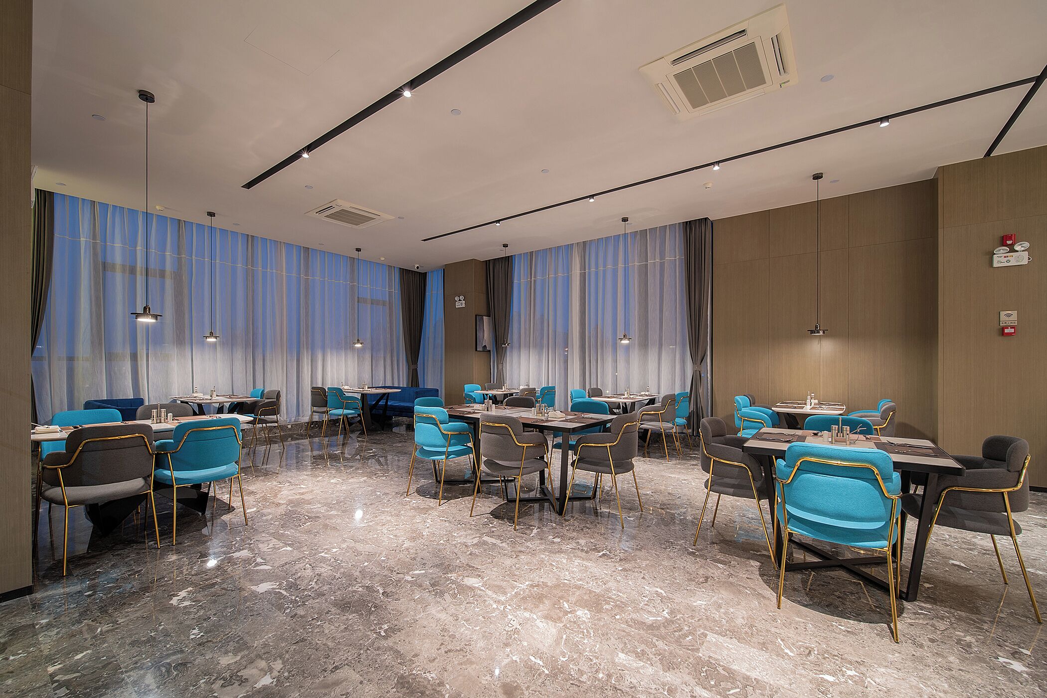 A hotel conference room with tables and chairs, large windows with curtains, and a marble floor.