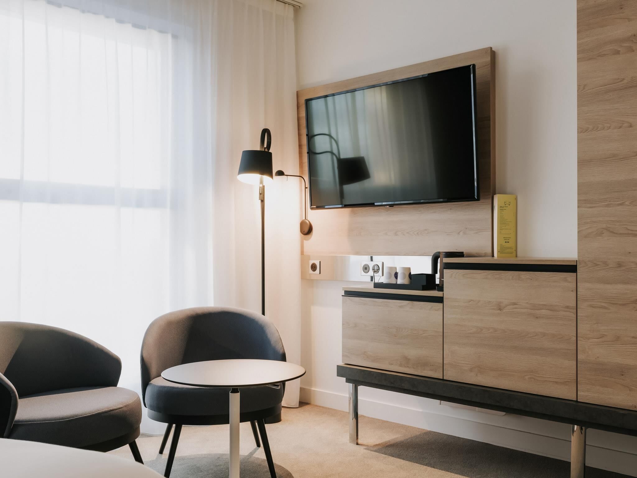 Quarto de hotel com televisão na parede, uma mesinha redonda, duas cadeiras, um armário e um abajur.
