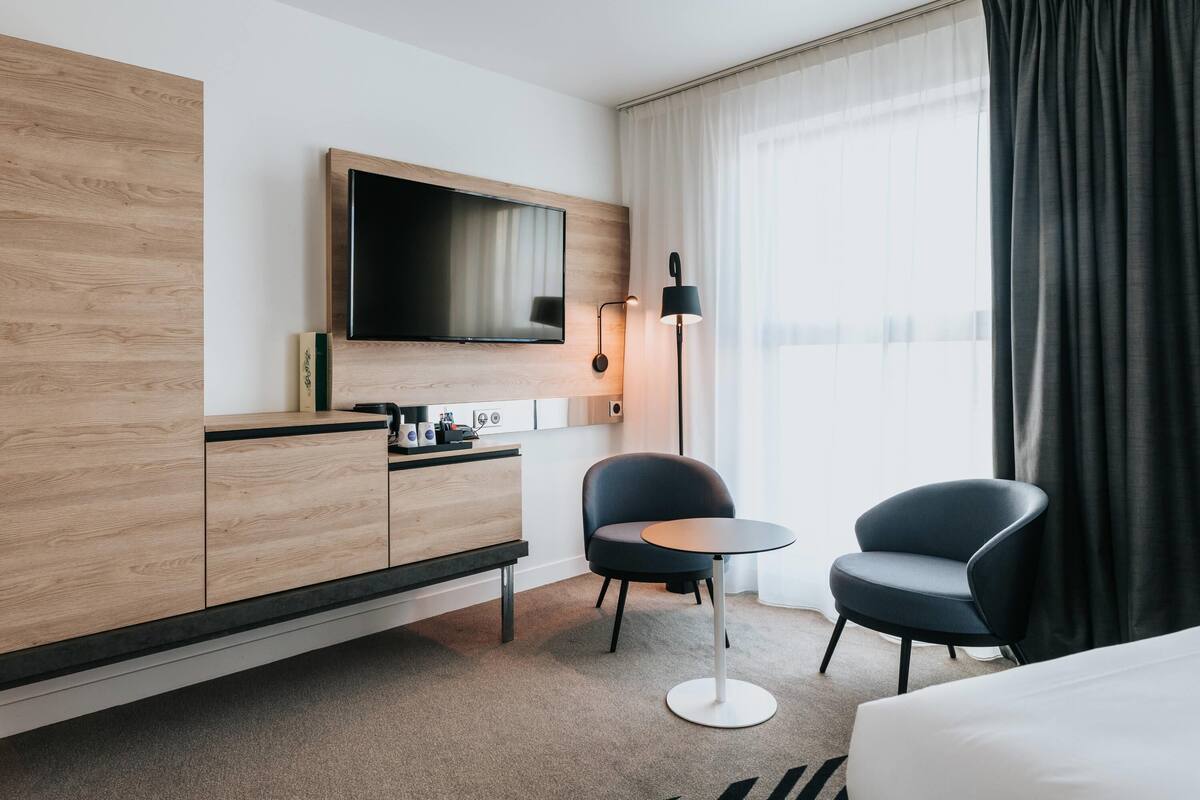 A modern hotel room with a flat-screen TV, a wooden cabinet, a small round table, two chairs, and a large window with curtains.