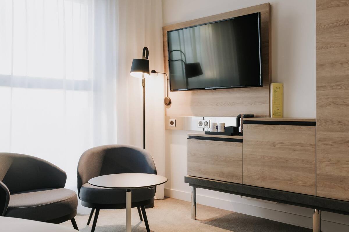 A hotel room with a flat-screen TV mounted on the wall, a small round table, two chairs, a cabinet with a wooden finish, and a floor lamp.