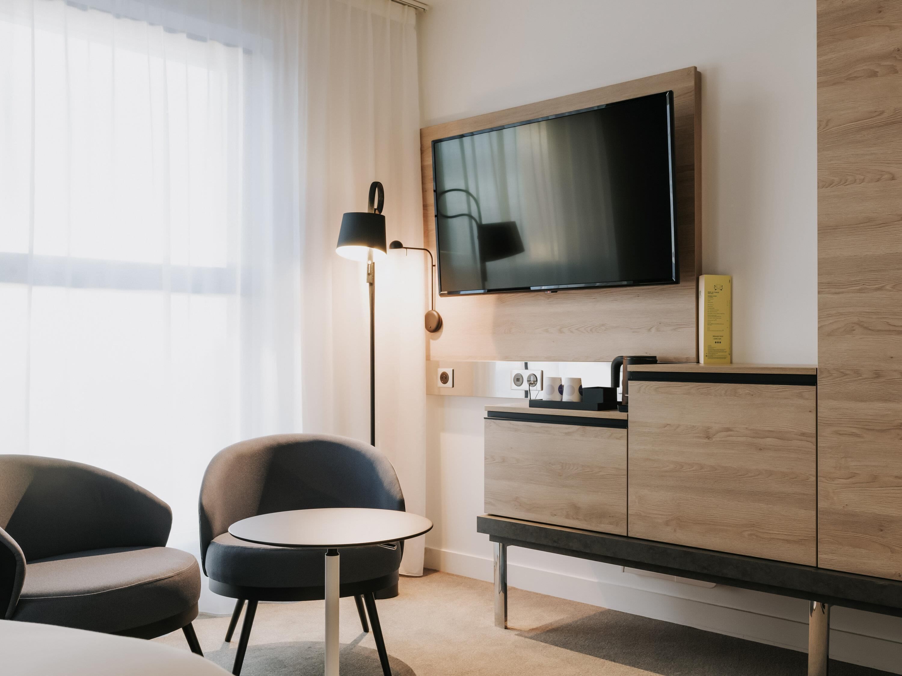 Ein Hotelzimmer mit einem an der Wand befestigten Flachbildfernseher, einem kleinen runden Tisch, zwei Stühlen, einer Holzkabine und einer Stehlampe.