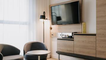 Ein Hotelzimmer mit einem an der Wand befestigten Flachbildfernseher, einem kleinen runden Tisch, zwei Stühlen, einer Holzkabine und einer Stehlampe.