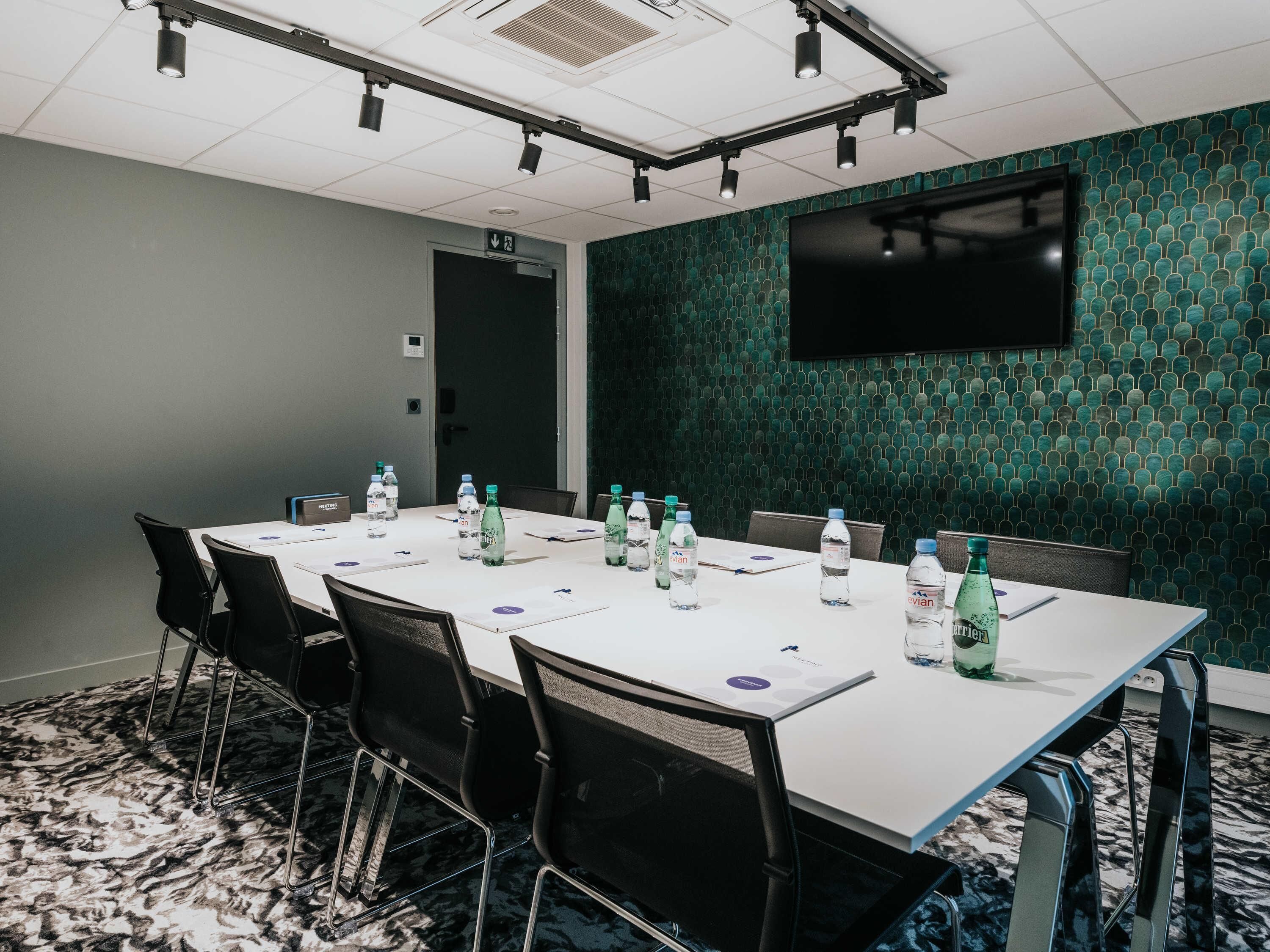 Une salle de conférence avec une longue table, des chaises, des bouteilles d’eau, un téléviseur à écran plat et un mur décoré de motifs.