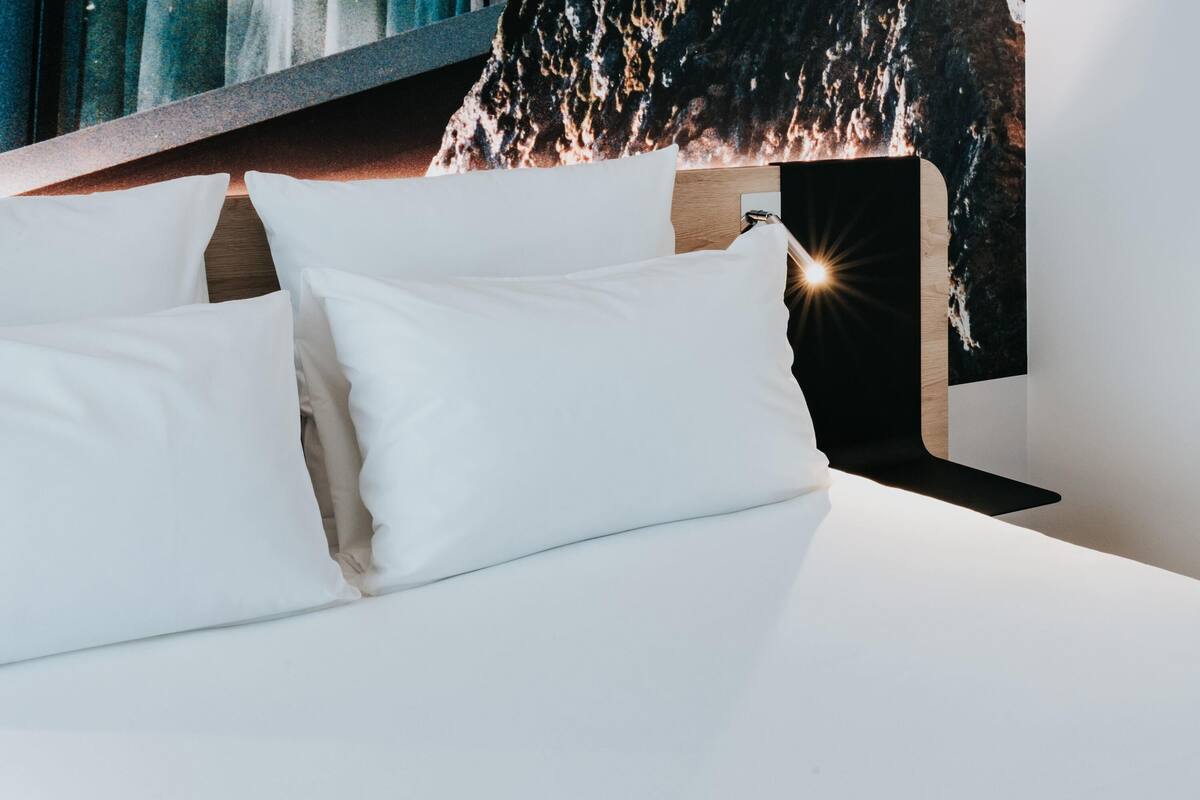 A neatly made bed with white linens and pillows, a wooden headboard, and a wall with a textured, abstract mural.