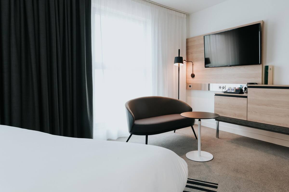 A modern hotel room with a large bed, a desk, a chair, a flat-screen TV, and a large window with sheer curtains.
