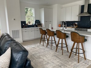Cocina moderna con armarios blancos, encimera negra y ventana con vistas a árboles. Hay tres taburetes de bar de madera y un sofá de cuero negro con un cojín decorado.