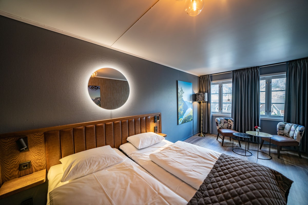 Ein Hotelzimmer mit einem großen Bett, einem runden Spiegel, einer Sitzecke mit einem Tisch und Blick ins Freie durch die Fenster.