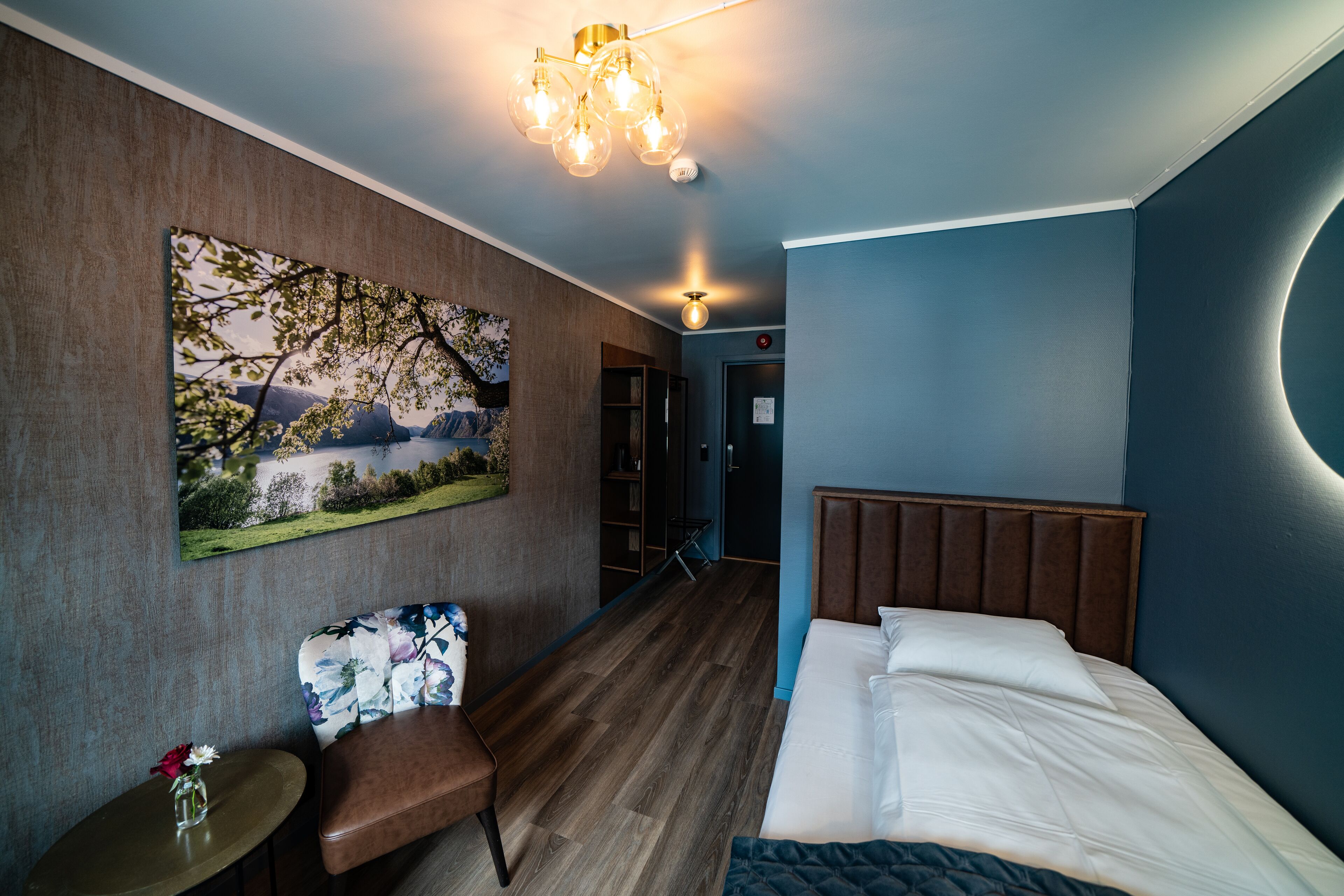 Quarto de hotel com cama, área de estar com uma cadeira e mesa, um quadro na parede e um lustre.