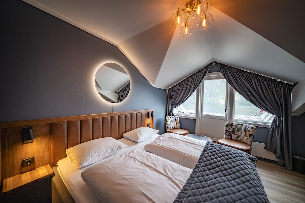 Ein modernes Hotelzimmer mit einem großen Bett, einem runden Spiegel, einem Nachttisch, einer Sitzecke mit einem Sessel und einem Fenster mit Vorhängen.