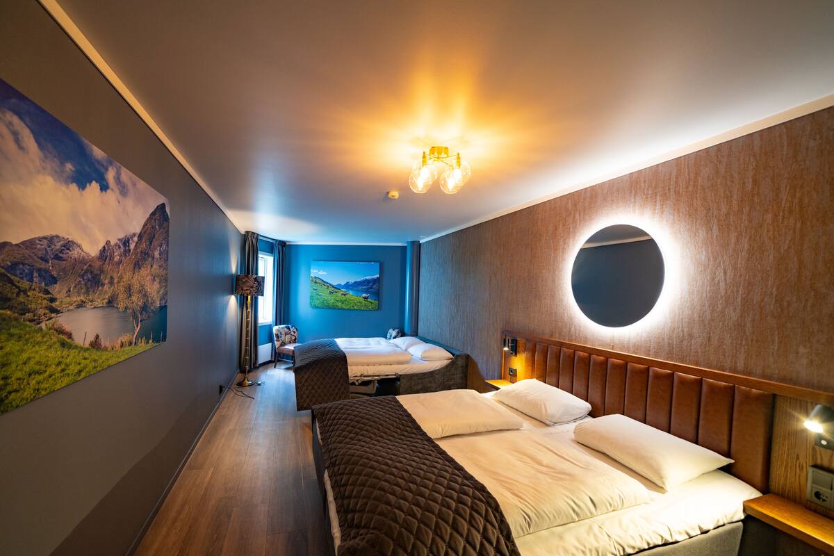 Ein modernes Hotelzimmer mit einem großen Bett, einem kleineren Bett, einem runden Spiegel und einem Gemälde einer Berglandschaft.