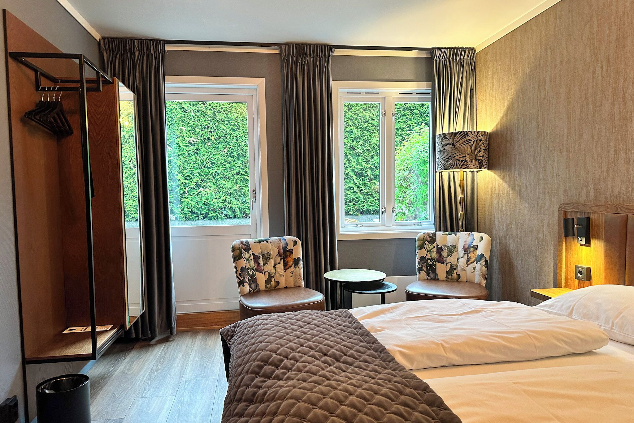 A hotel room with a bed, two chairs, a small table, a wardrobe, and a window with curtains.