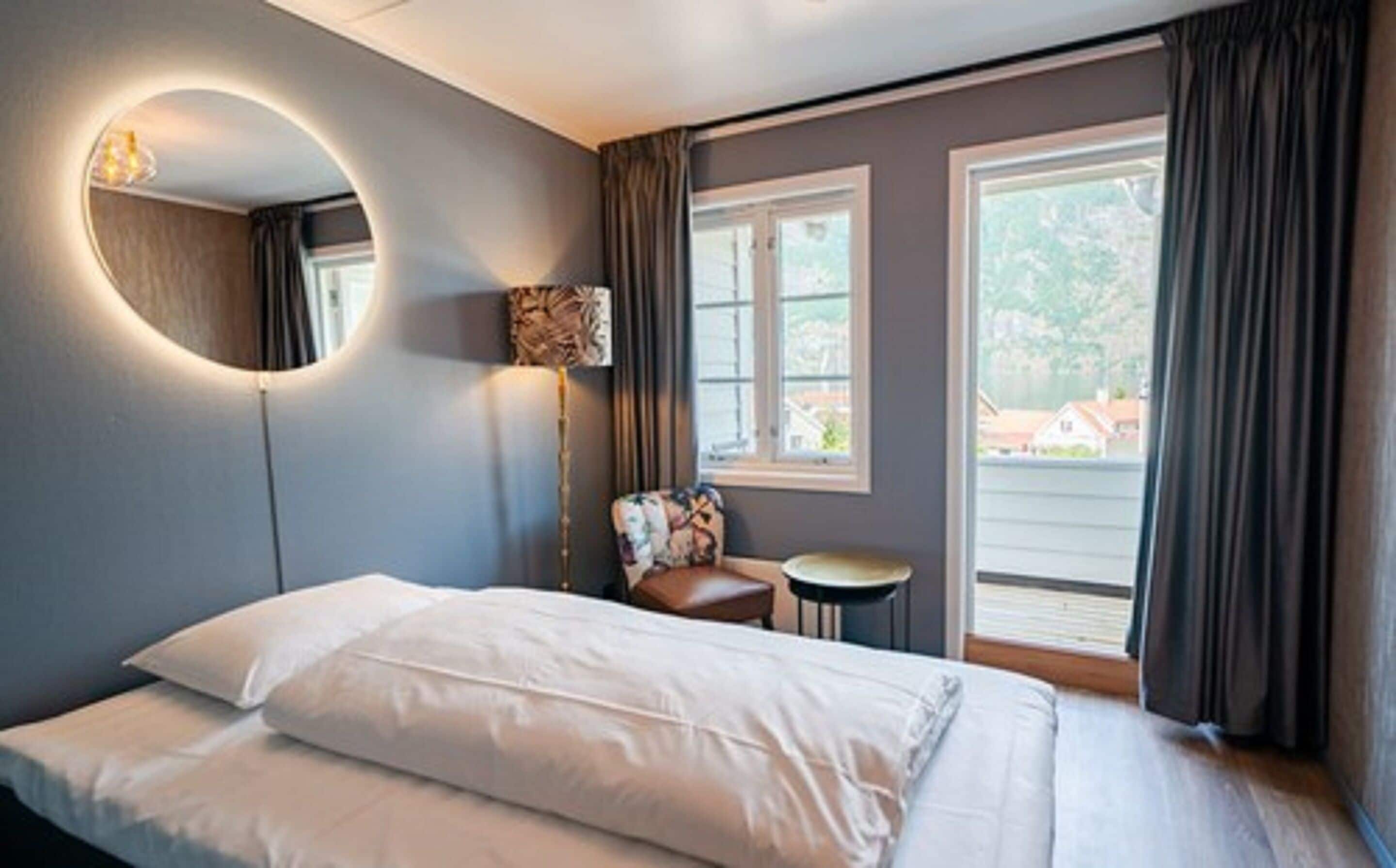 Une chambre d’hôtel avec un lit, un miroir rond, une petite table, une chaise et une fenêtre avec des rideaux.