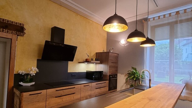 Eine moderne Küche mit Holzausstattung, einer zentralen Kochinsel, Pendelleuchten und Blick ins Freie durch große Fenster.