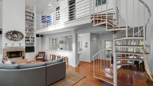 A spacious living area with a high ceiling, a fireplace, a large gray sofa, and a staircase leading to an upper level.