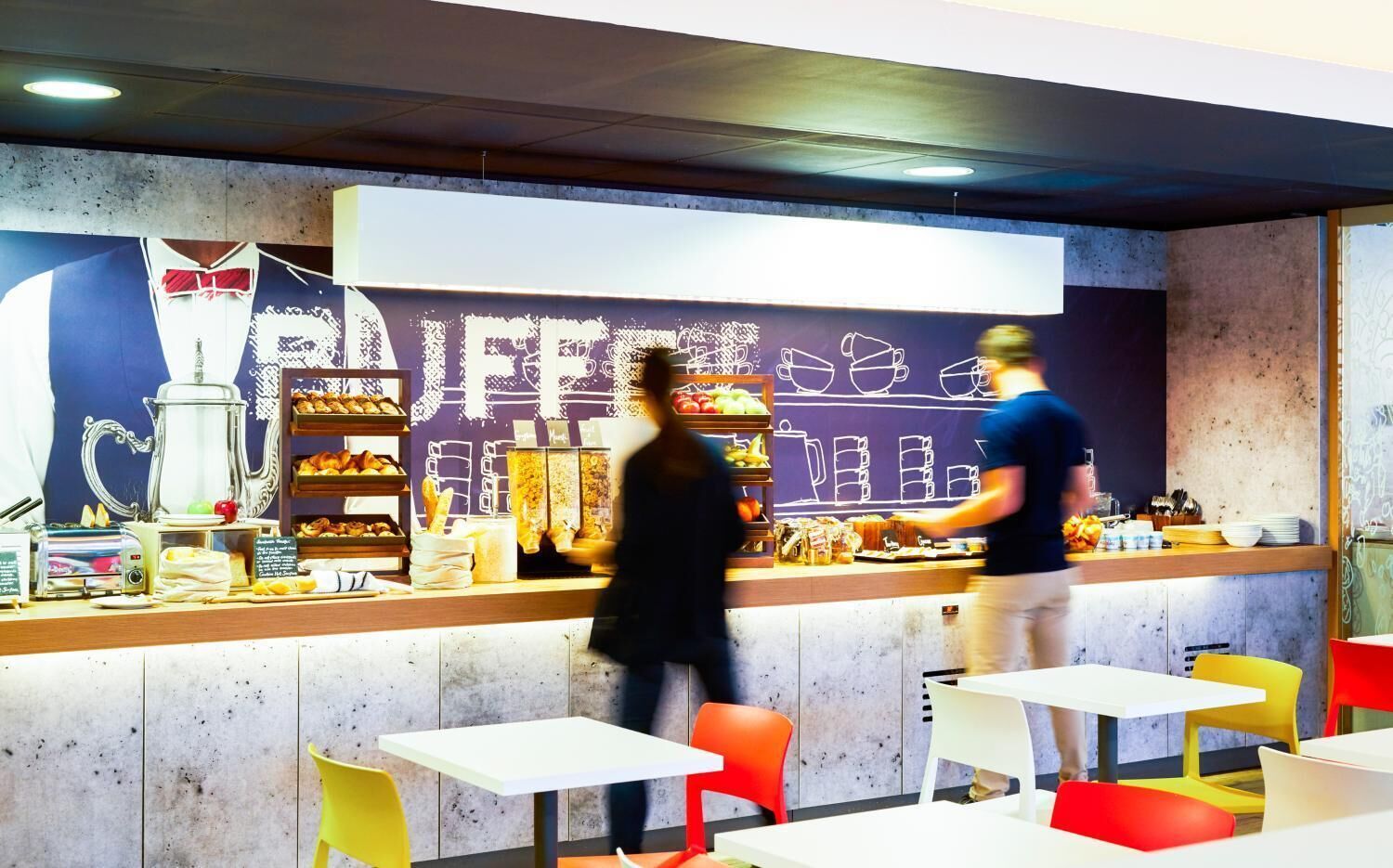 Un café avec un comptoir où sont exposées des pâtisseries et du matériel pour préparer le café, des tables avec des chaises, et un mur orné d’une fresque sur le thème du café.