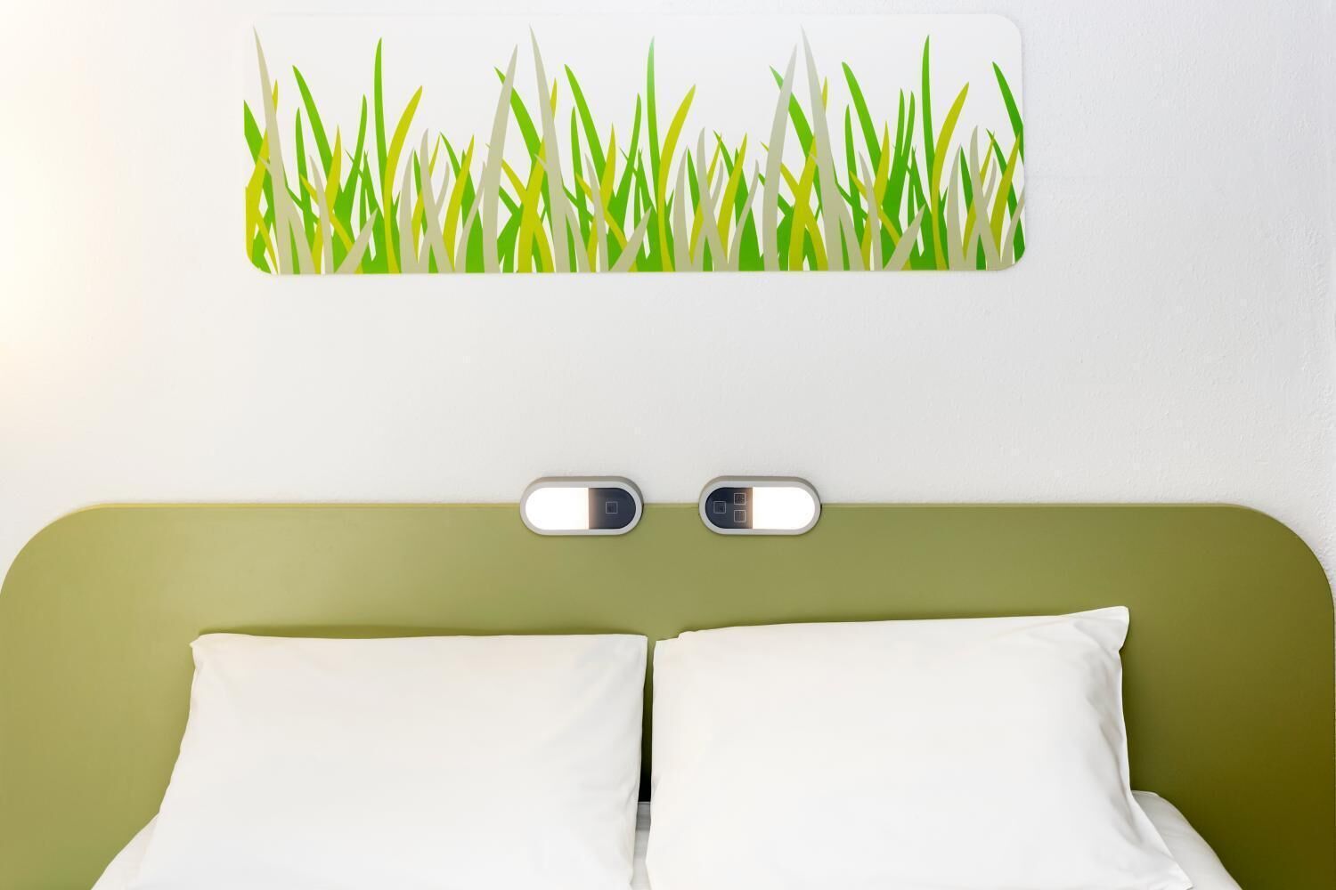 A hotel room with a green headboard, two white pillows, and a wall-mounted light fixture.