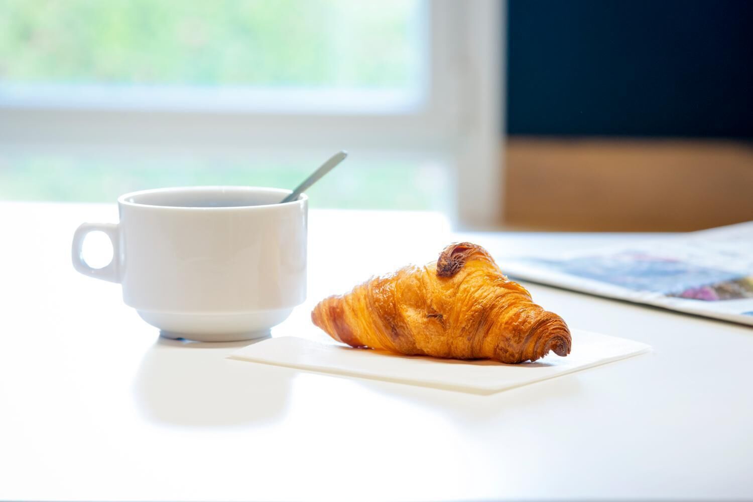 Un croissant sur une assiette blanche, à côté d’une tasse blanche avec une cuillère, sur une table où se trouve un magazine.