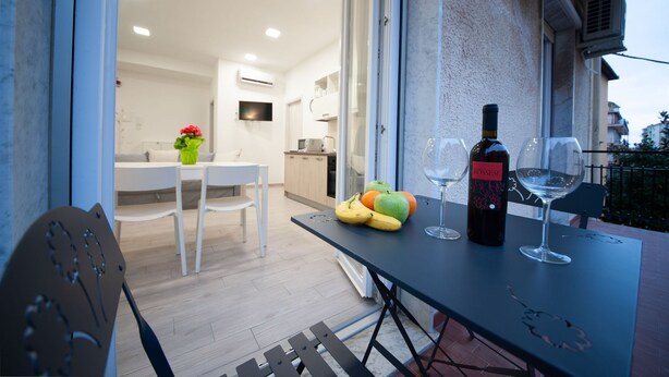 A modern kitchen with white cabinets, a dining area with a table and chairs, and a balcony with a table set for two with wine glasses and fruit.