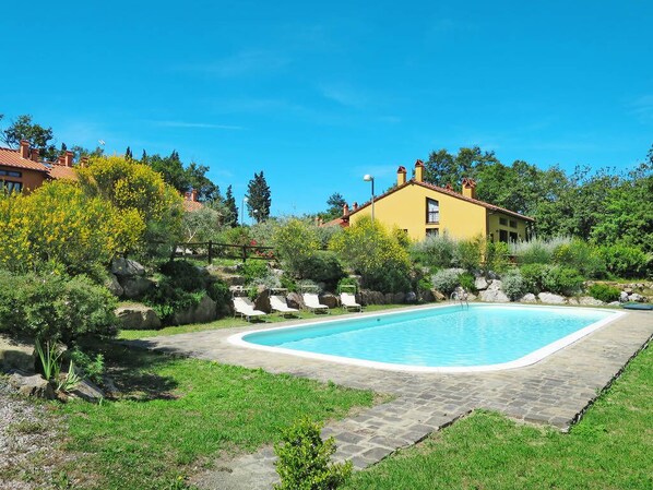 A swimming pool with lounge chairs, surrounded by greenery and a yellow building in the background.