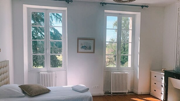 Ein Zimmer mit zwei Betten, einem Heizkörper, einem Schminktisch und großen Fenstern mit Blick auf Bäume.