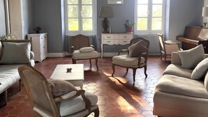 Ein Zimmer mit einer Couch, Sesseln, einem Tisch, einem Klavier und großen Fenstern mit Vorhängen.