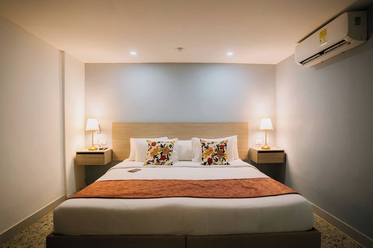 Een hotelkamer met een groot bed, twee bedlampen, een houten hoofdbord en een airconditioner aan de muur.