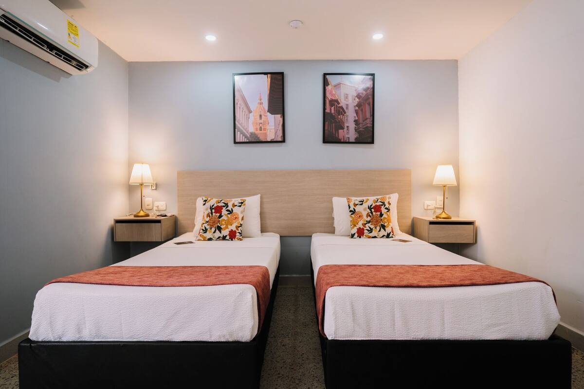 Een hotelkamer met twee bedden, een houten hoofdbord, twee nachtkastjes met lampen en twee ingelijste foto's aan de muur.