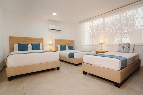 A hotel room with three beds, each with a wooden headboard, white bedding, and blue accents. There is a window with a lattice design, and an air conditioning unit mounted on the wall. - Casa Franco Hotel (Cartagena)