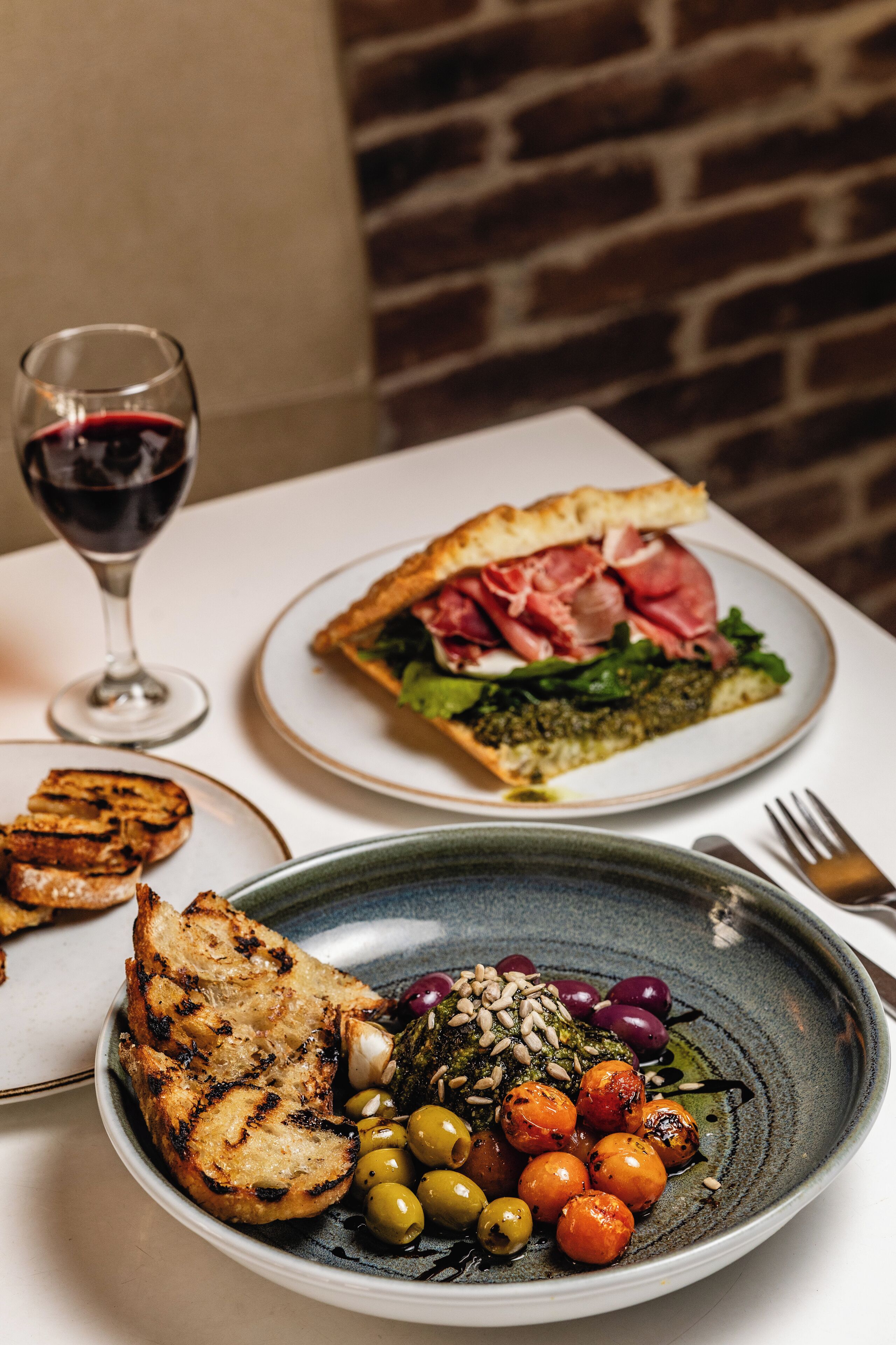 A table with a plate of grilled bread, a plate with a sandwich, and a glass of red wine.