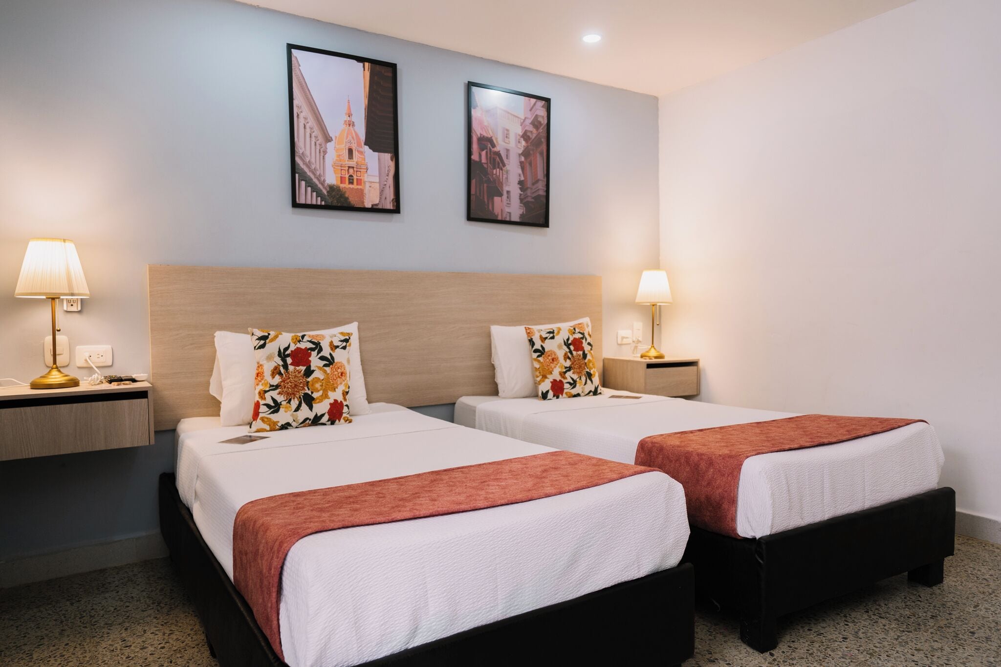 A hotel room with two beds, each with a wooden headboard, white bedding, and a red throw blanket. There are two bedside tables with lamps, and the walls are adorned with framed pictures.