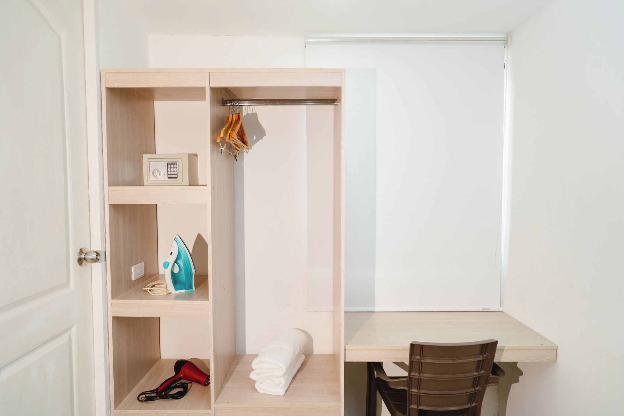A compact room with a wooden desk, a chair, a wardrobe with shelves, a hanging rack, and a small safe.