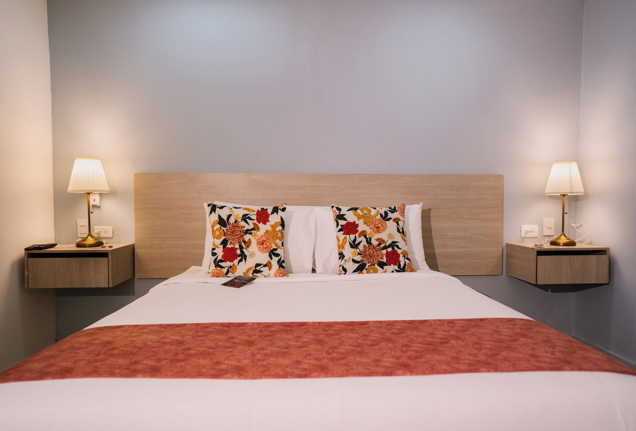 A neatly made bed with floral pillows, a red blanket, and bedside lamps on wooden nightstands.